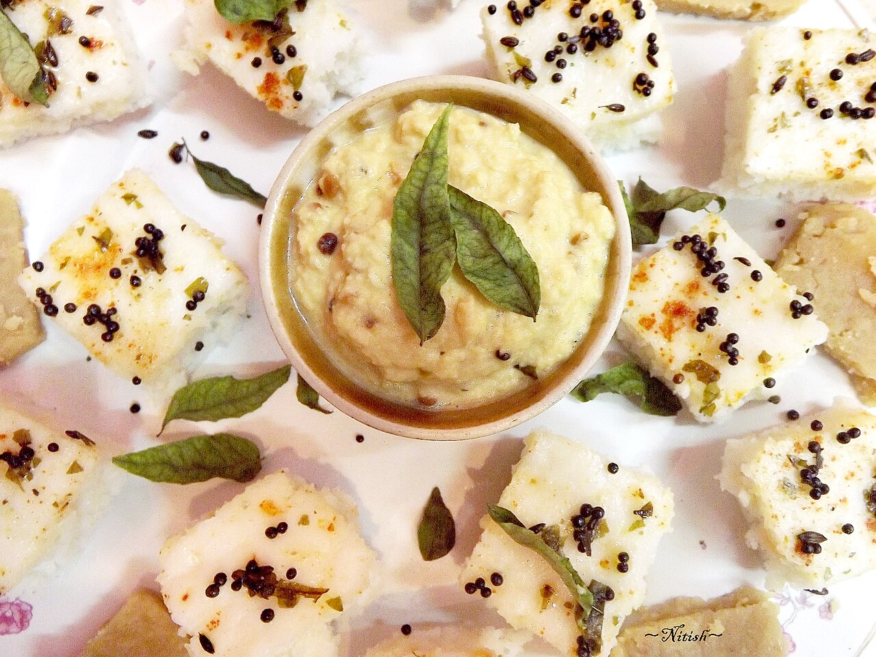 Squares of white dhokla tempered with mustard seeds and curry leaves, around a bowl of chutney