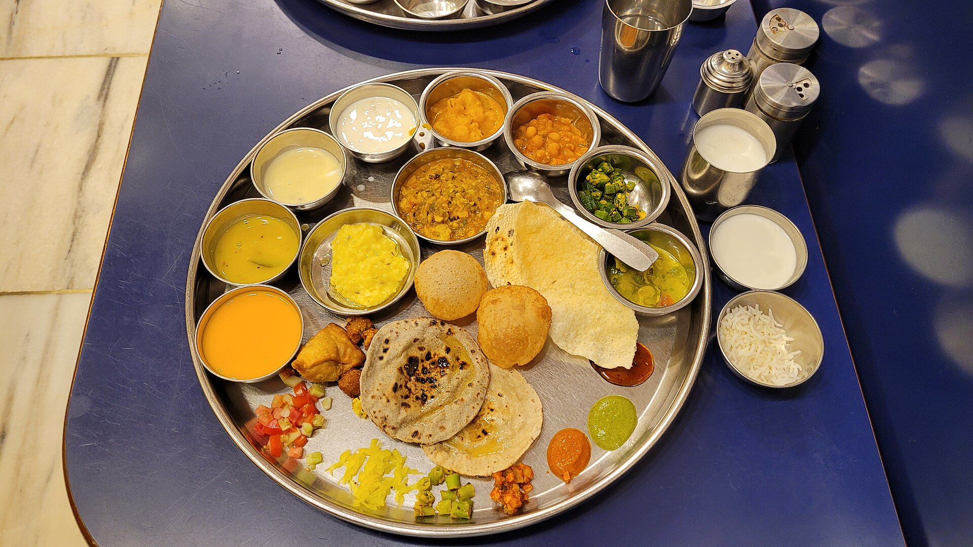 A full steel thali from above: dal, kadhi, three shaaks, farsan, puri, rotli, papad and sweets in small katoris